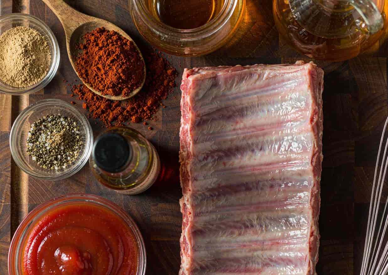 Overhead flat lay of barbecue rib ingredients including a rack of ribs, smoked paprika, and brown sugar