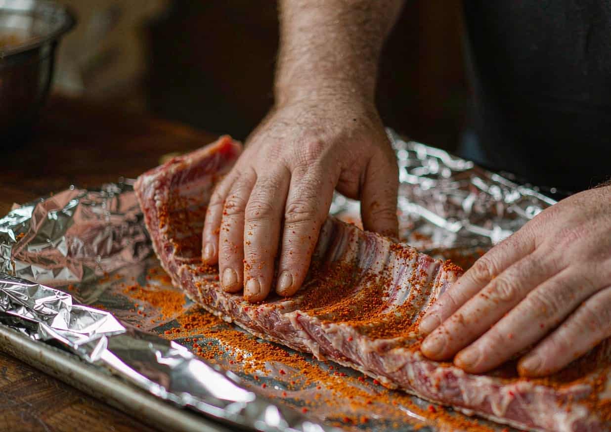 A rack of pork ribs being coated with dry spice rub on aluminium foil