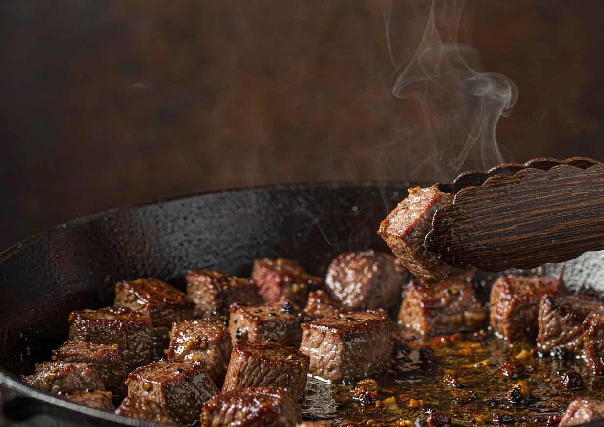Beef chuck cubes browning in a cast iron Dutch oven with tongs lifting one cube