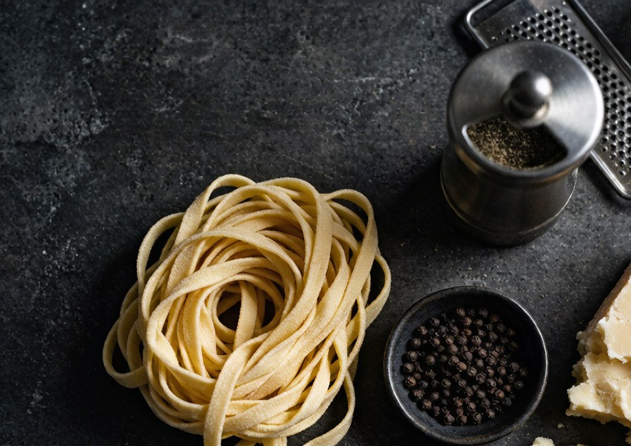 Overhead flat lay of tonnarelli pasta, a wedge of Pecorino Romano, black peppercorns and a microplane