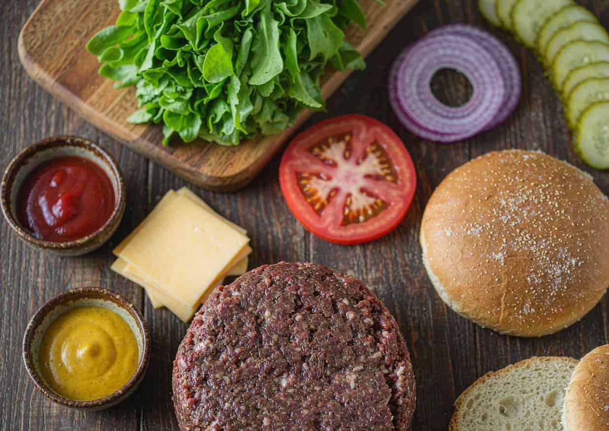 Overhead flat lay of cheeseburger ingredients including ground chuck, potato buns, cheese, and vegetables