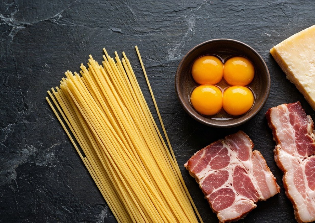 Overhead flat lay of spaghetti, guanciale, egg yolks, Pecorino Romano and black peppercorns