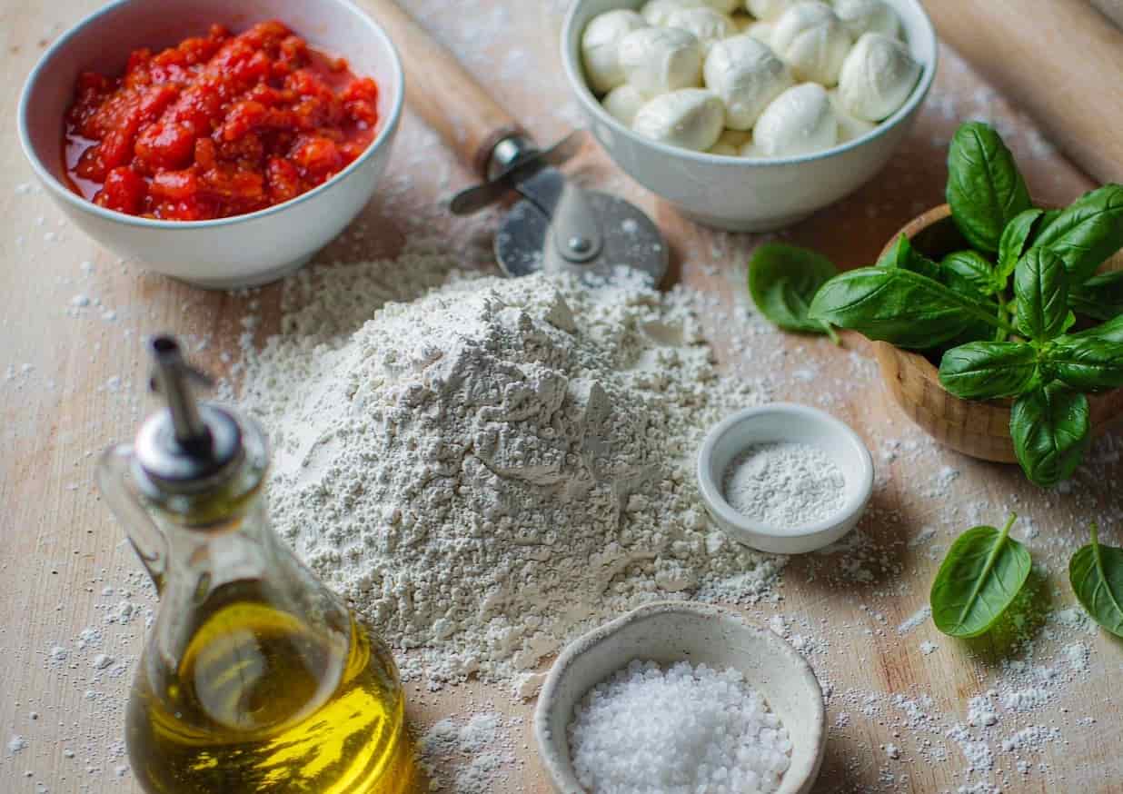 Overhead flat lay of pizza ingredients including 00 flour, San Marzano tomatoes, mozzarella, and basil