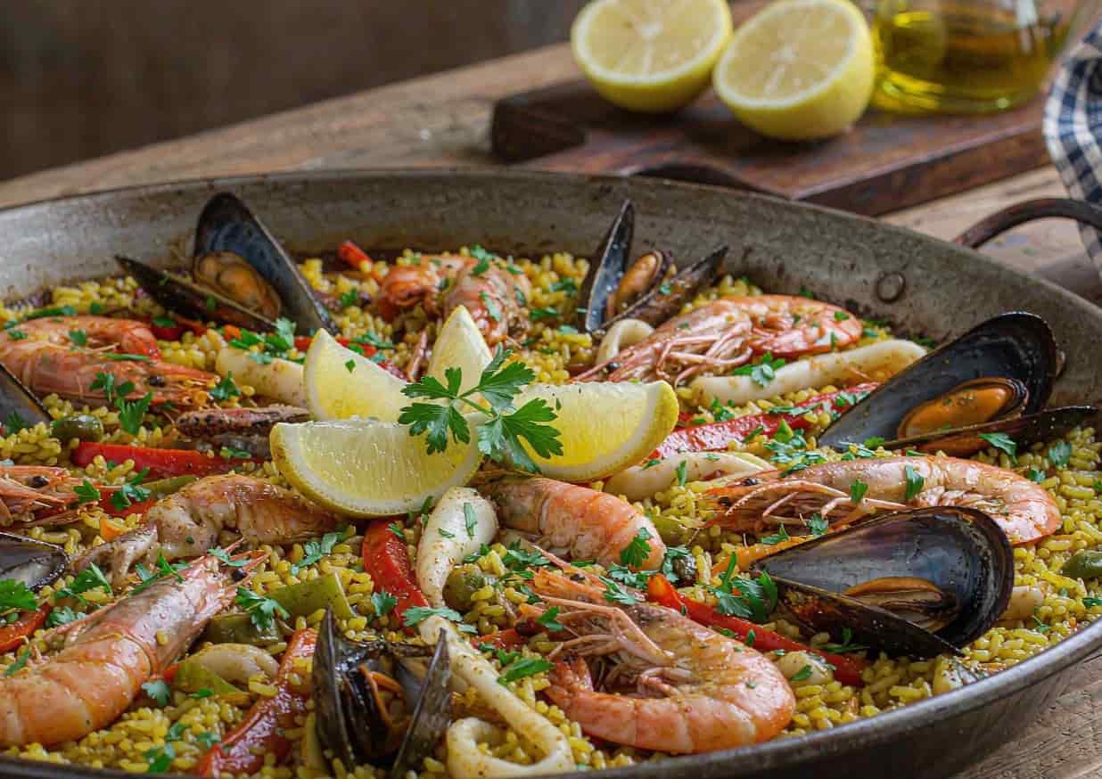 A shallow pan of Spanish seafood paella with shrimp, mussels, calamari, and lemon wedges