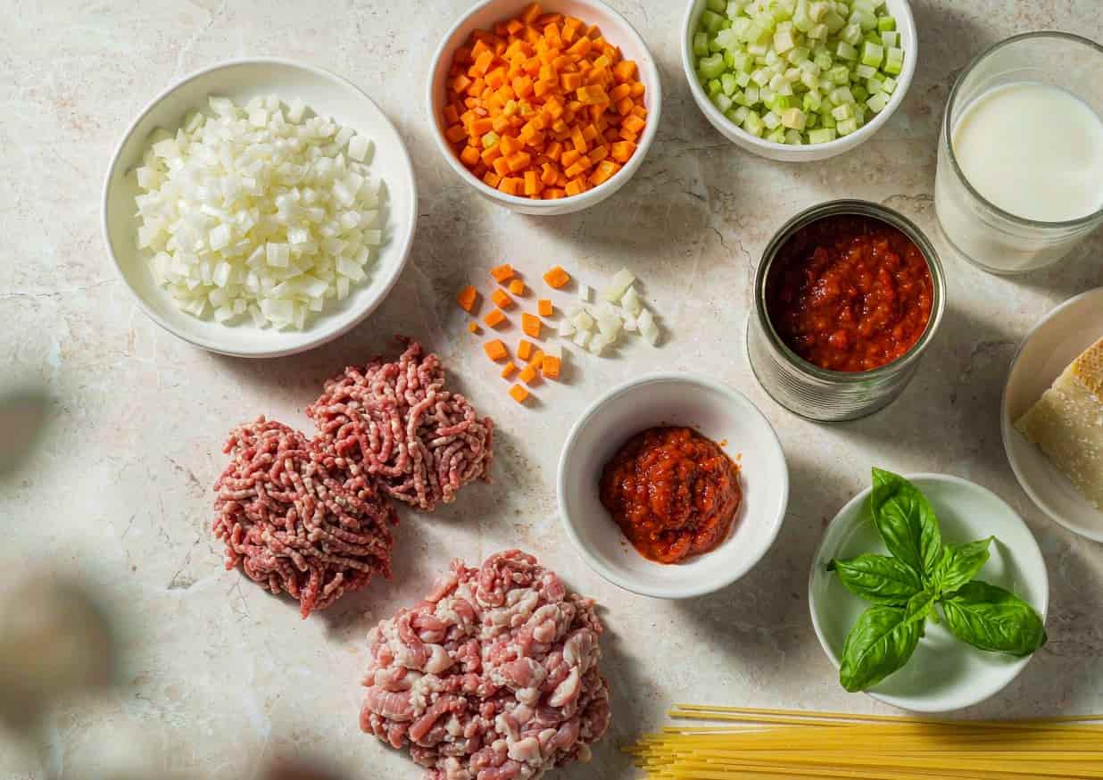 Overhead flat lay of spaghetti Bolognese ingredients including soffritto, ground beef, and canned tomatoes
