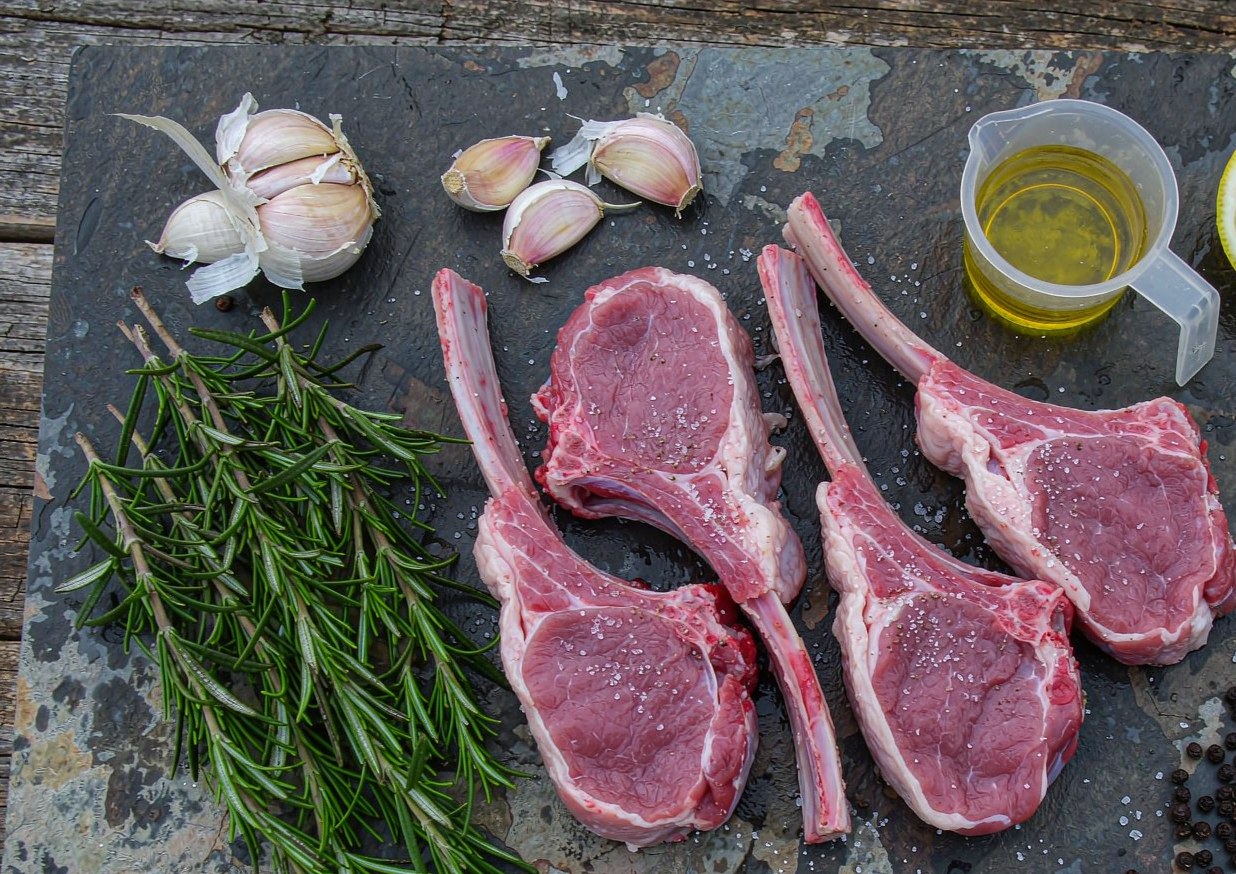 Overhead flat lay of lamb chops with garlic cloves, fresh rosemary, lemons, olive oil and black pepper