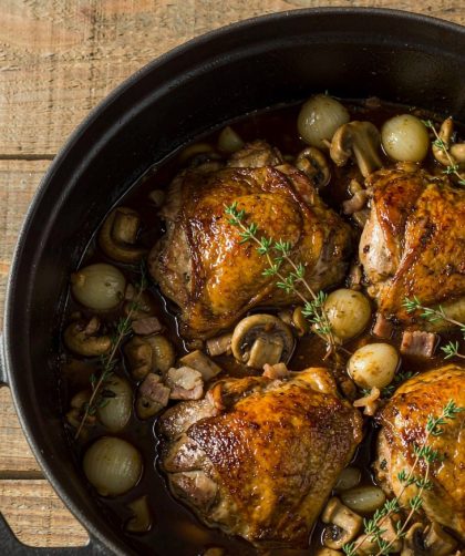 Coq au vin in a cast-iron casserole with browned chicken pieces, mushrooms and pearl onions in a glossy red wine sauce