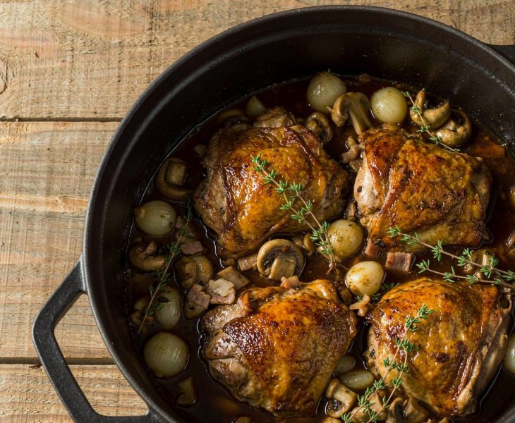Coq au vin in a cast-iron casserole with browned chicken pieces, mushrooms and pearl onions in a glossy red wine sauce