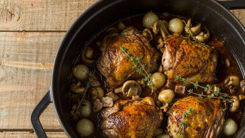 Coq au vin in a cast-iron casserole with browned chicken pieces, mushrooms and pearl onions in a glossy red wine sauce