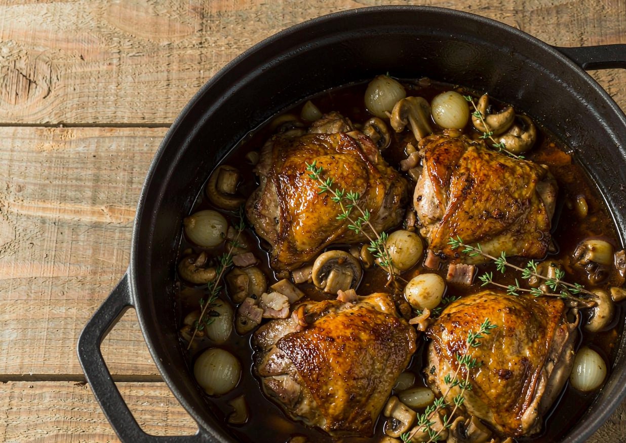 Coq au vin in a cast-iron casserole with browned chicken pieces, mushrooms and pearl onions in a glossy red wine sauce