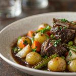French lamb navarin in a wide shallow bowl, chunks of browned lamb with glazed new potatoes, carrots, turnips, peas and green beans in a glossy dark sauce scattered with chopped parsley