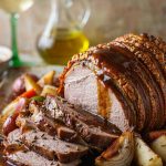 Sliced German pork roast with a crackled brown crust on a platter with pan gravy, roasted root vegetables, and caraway seeds