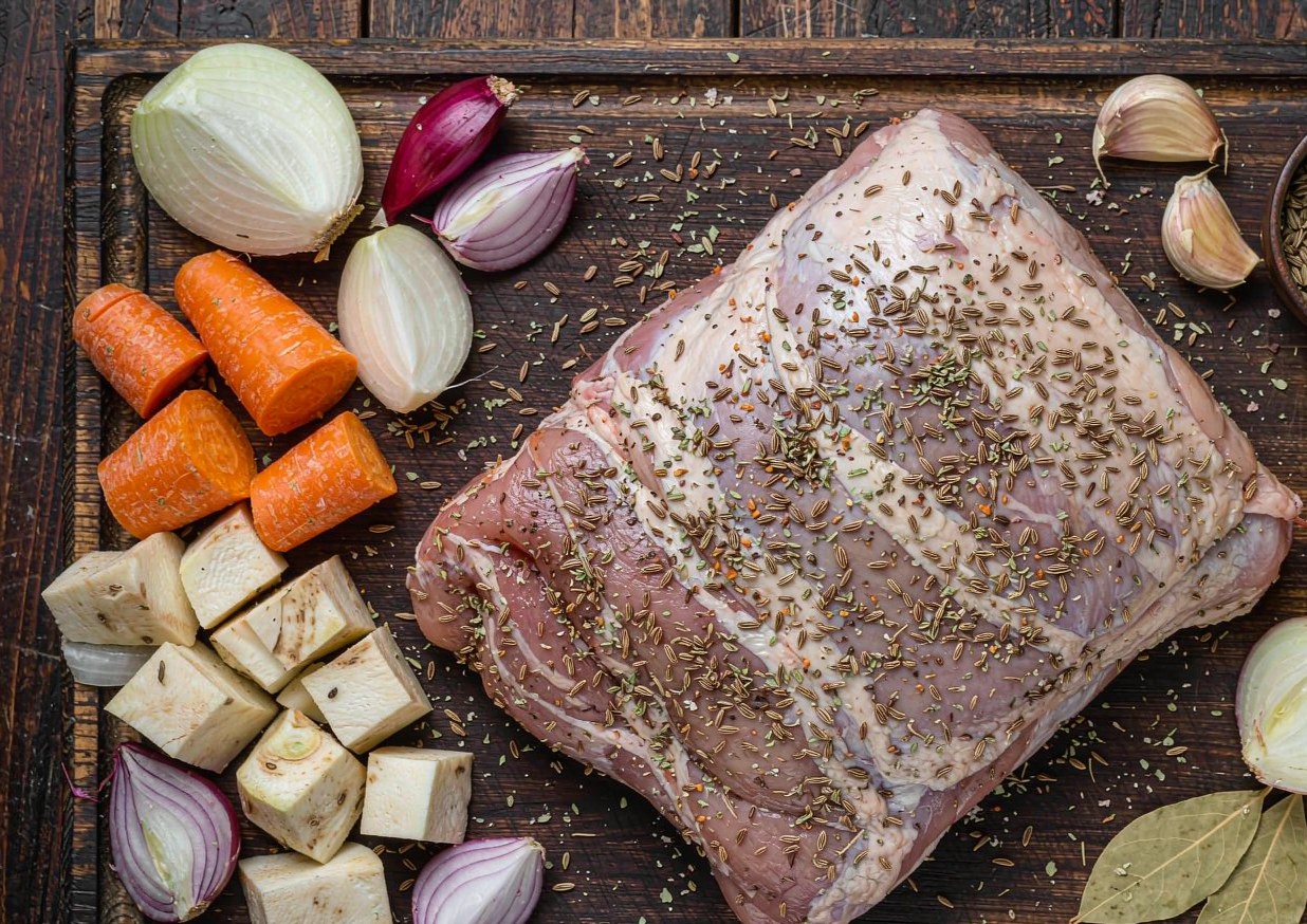 Overhead flat lay of pork shoulder, caraway seeds, dried marjoram, garlic, onions, carrots, celeriac, and bay leaves on a dark wooden board