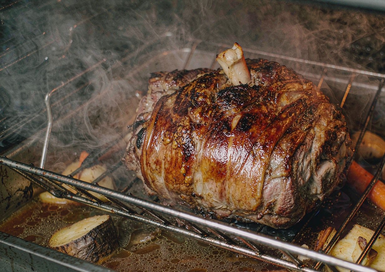 Pork shoulder roasting on a rack in a heavy roasting pan surrounded by onions and root vegetables in bubbling stock