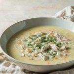 Bowl of pale gold Greek avgolemono soup with rice, shredded chicken, carrot, and chopped dill, with lemon halves beside it