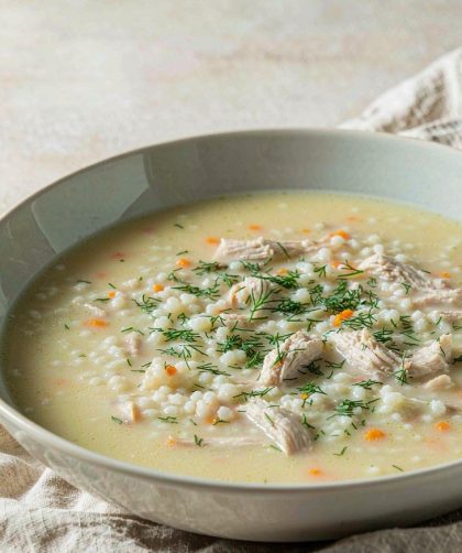 Bowl of pale gold Greek avgolemono soup with rice, shredded chicken, carrot, and chopped dill, with lemon halves beside it