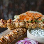 Grilled chicken souvlaki skewers on a platter with tzatziki, sliced red onion, and warm flatbread