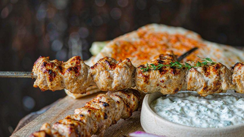 Grilled chicken souvlaki skewers on a platter with tzatziki, sliced red onion, and warm flatbread