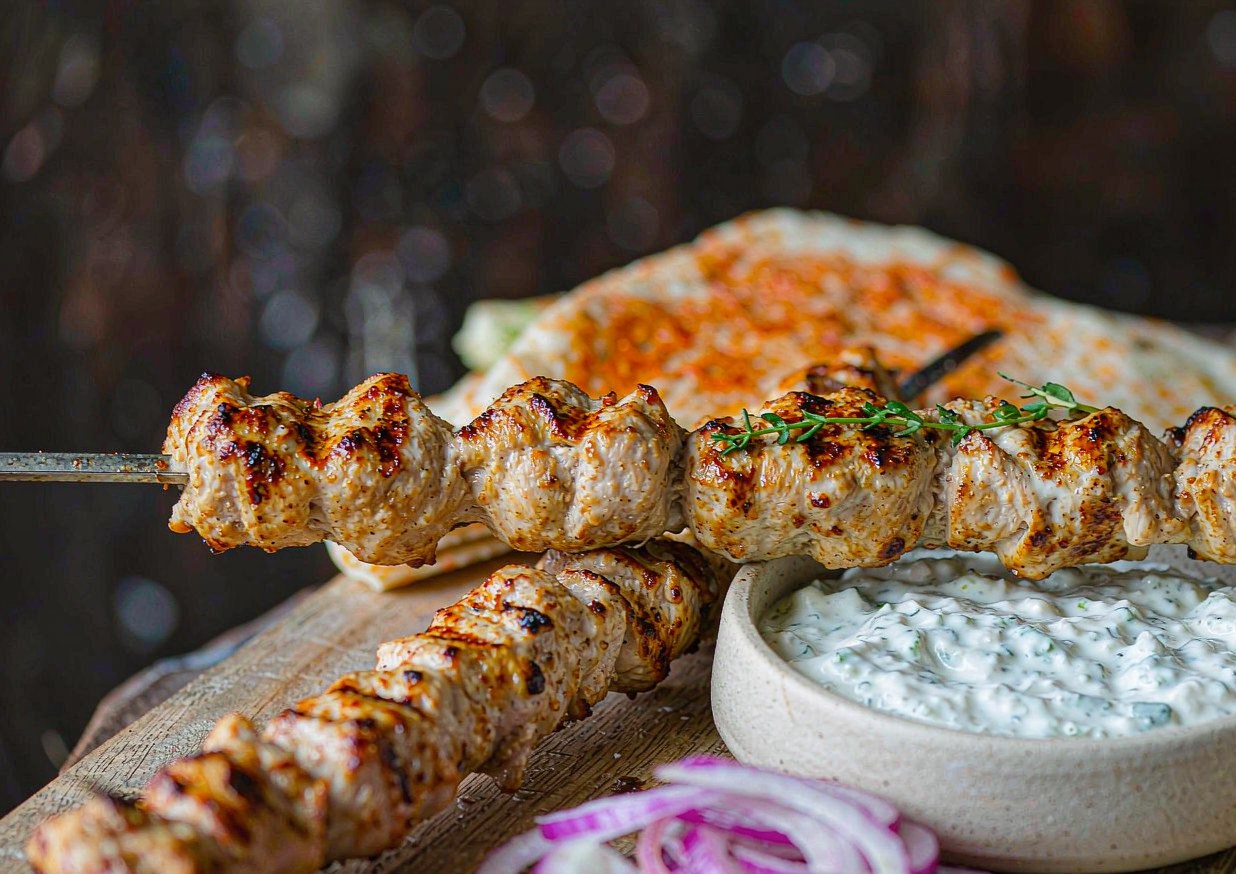 Grilled chicken souvlaki skewers on a platter with tzatziki, sliced red onion, and warm flatbread