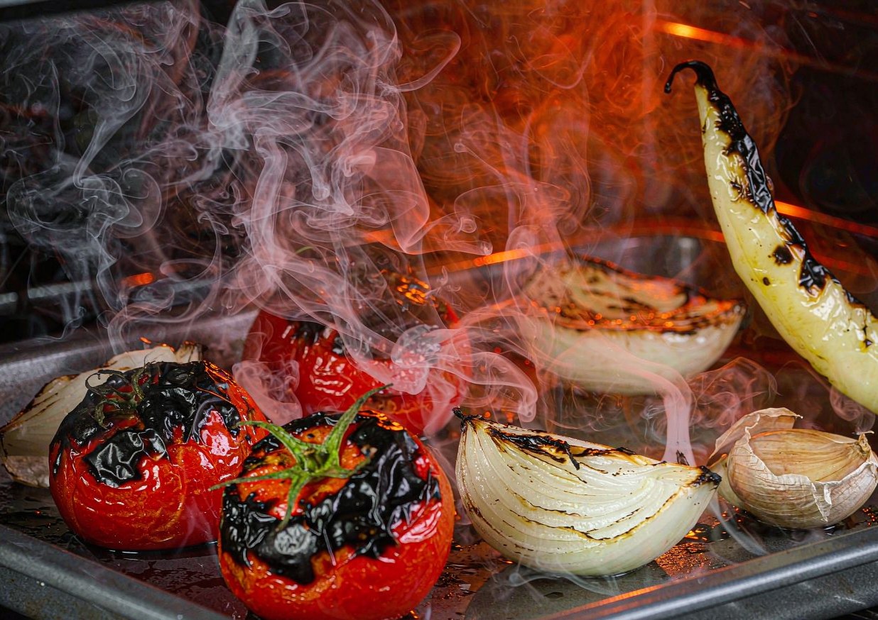 Charred tomatoes, onion, garlic and serrano chilli blistering under a broiler on a rimmed sheet pan with blackened spots on the skins