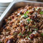 Shredded pork carnitas crisp and caramelised on a rimmed sheet pan with charred edges, scattered with diced onion, cilantro, and lime wedges