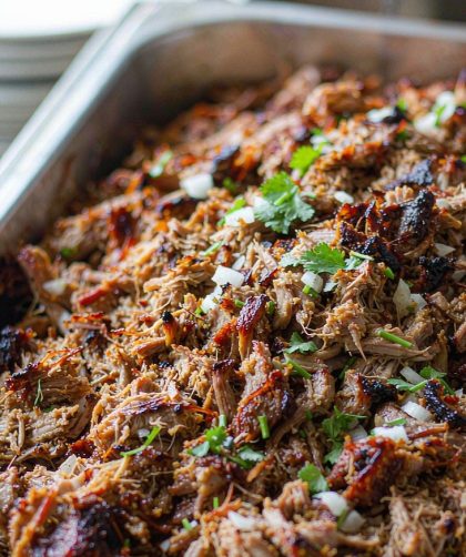 Shredded pork carnitas crisp and caramelised on a rimmed sheet pan with charred edges, scattered with diced onion, cilantro, and lime wedges