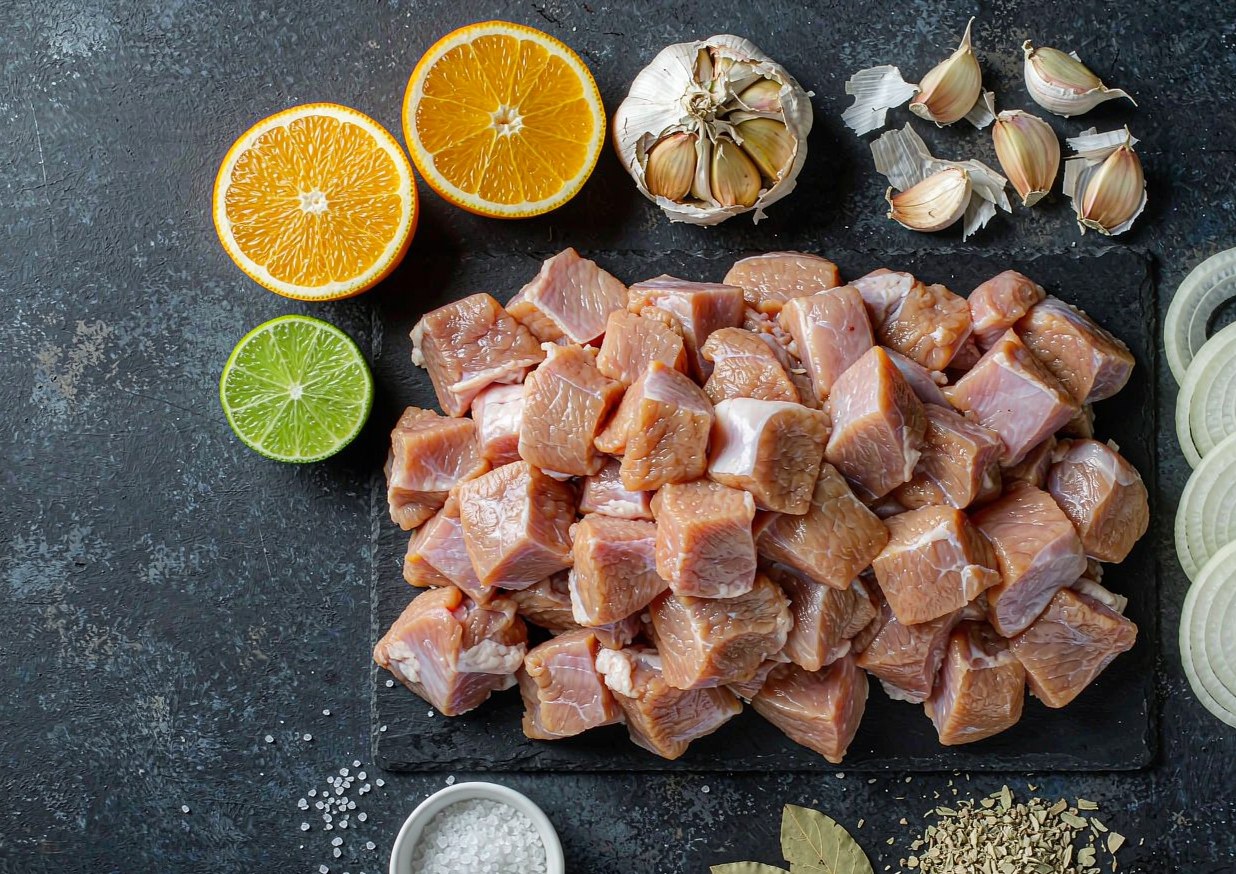 Overhead flat lay of cubed pork shoulder, oranges, limes, garlic, white onion, and dried Mexican oregano on a dark board