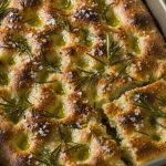 Golden dimpled rosemary focaccia in a rectangular metal baking tin with olive oil pools and flaky salt