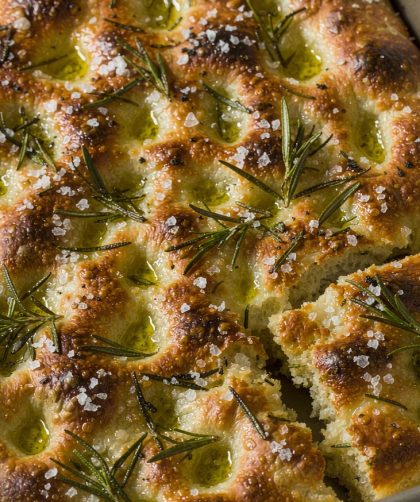 Golden dimpled rosemary focaccia in a rectangular metal baking tin with olive oil pools and flaky salt
