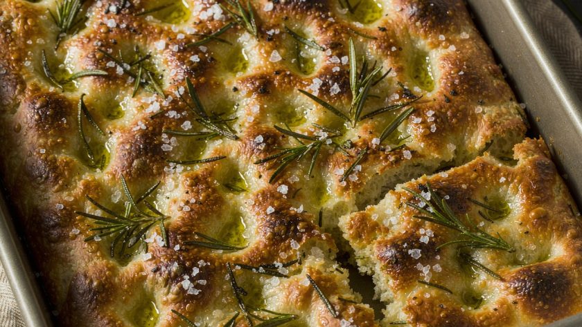 Golden dimpled rosemary focaccia in a rectangular metal baking tin with olive oil pools and flaky salt