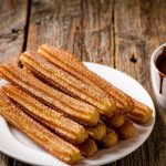 Golden ridged Spanish churros piled on a plate with cinnamon sugar beside a cup of thick hot chocolate for dipping