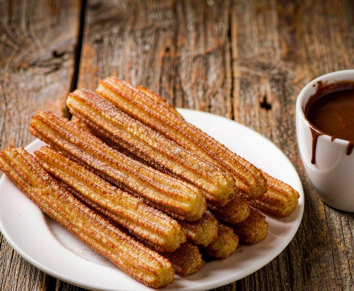 Golden ridged Spanish churros piled on a plate with cinnamon sugar beside a cup of thick hot chocolate for dipping