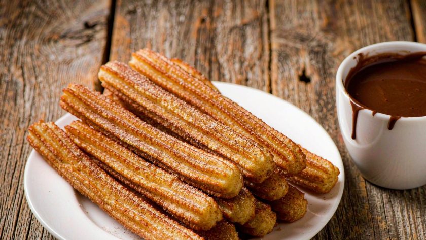 Golden ridged Spanish churros piled on a plate with cinnamon sugar beside a cup of thick hot chocolate for dipping
