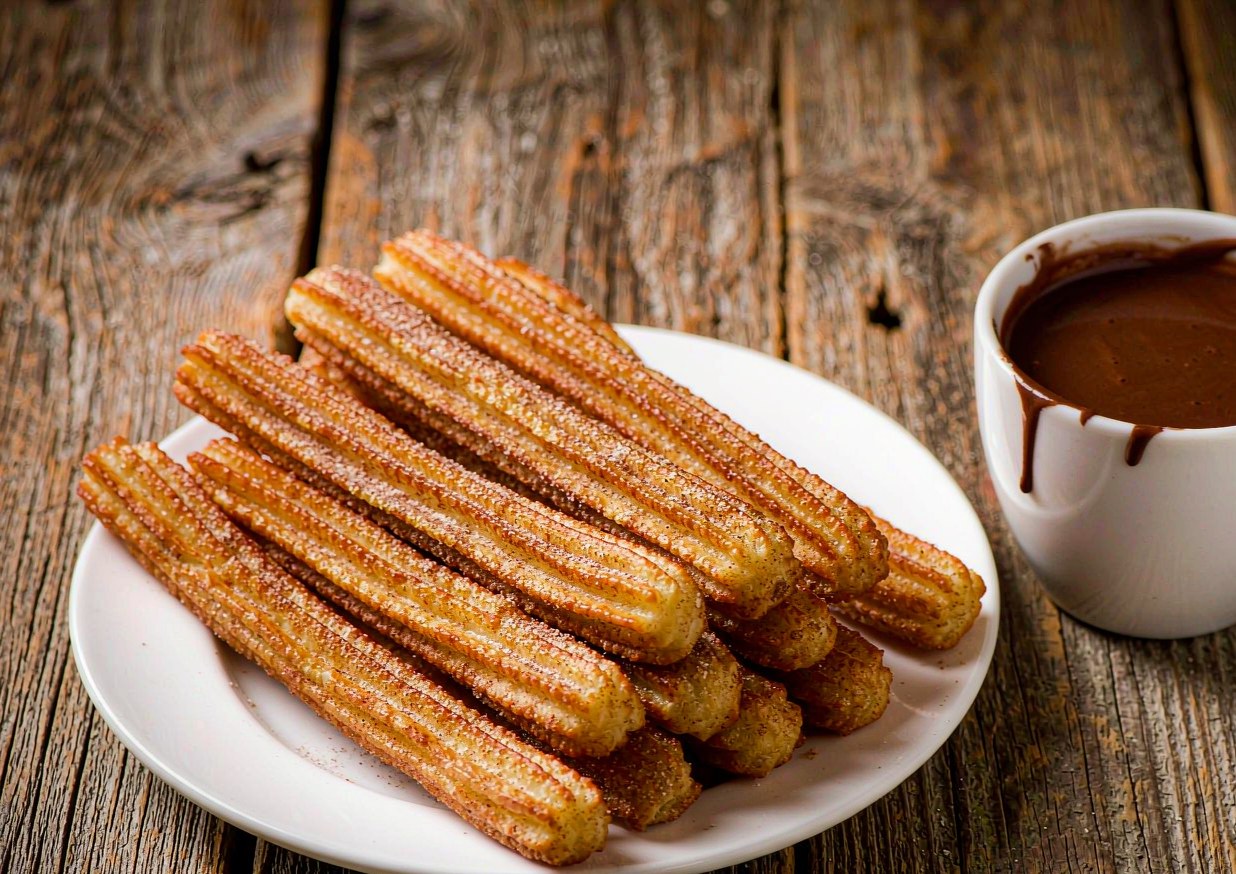 Golden ridged Spanish churros piled on a plate with cinnamon sugar beside a cup of thick hot chocolate for dipping