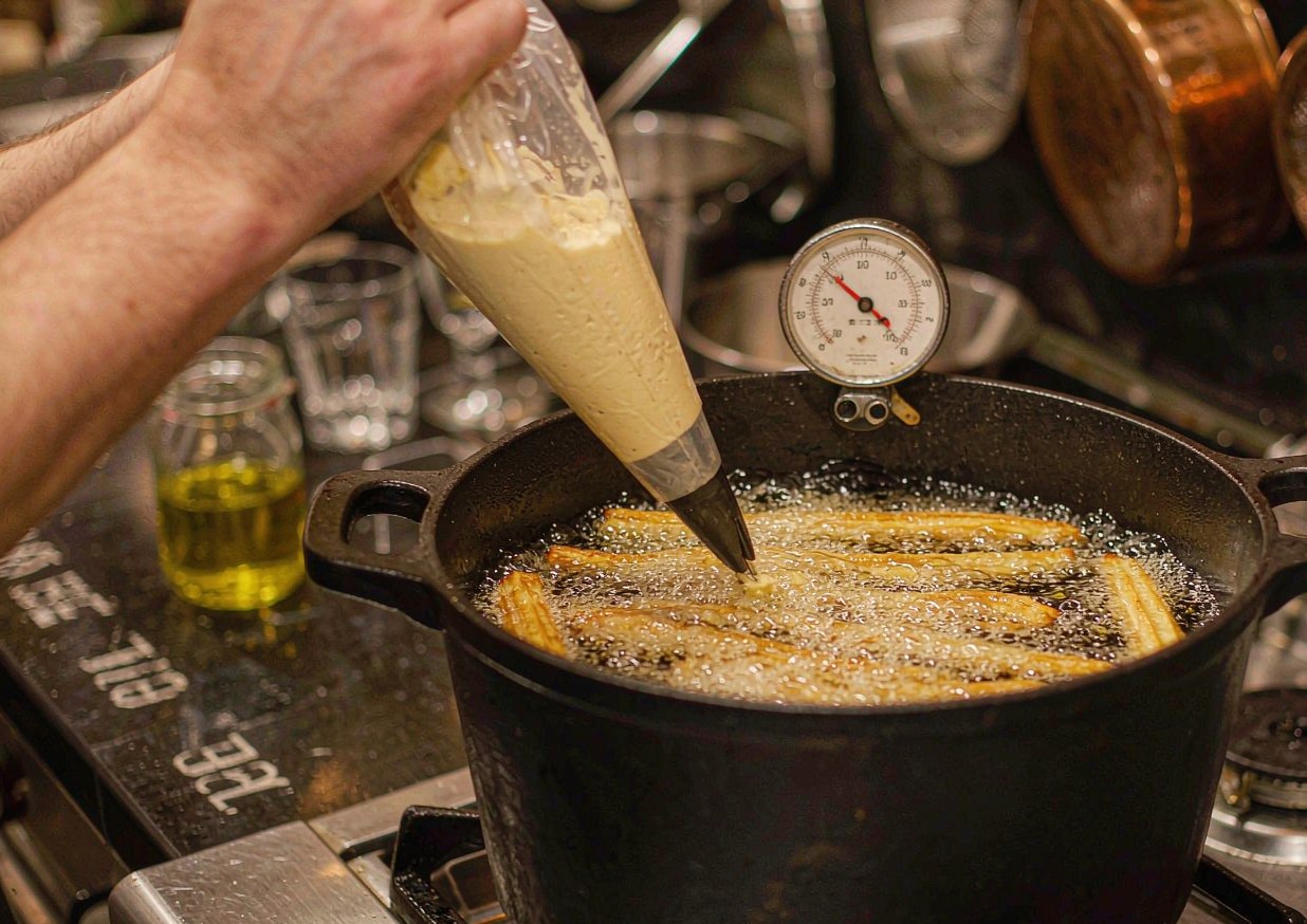 Churros dough being piped through a star nozzle directly into hot oil in a deep heavy pan
