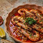 Charred garlic shrimp in a shallow terracotta dish glistening with red paprika oil, scattered with chopped parsley and lemon wedges