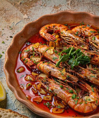Charred garlic shrimp in a shallow terracotta dish glistening with red paprika oil, scattered with chopped parsley and lemon wedges