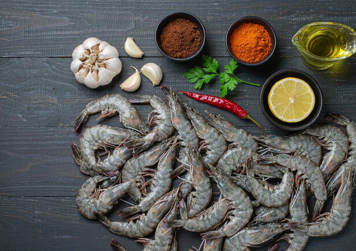 Overhead flat lay of large raw shrimp, a head of garlic, smoked paprika, dried chilli, parsley, and a lemon on a dark surface