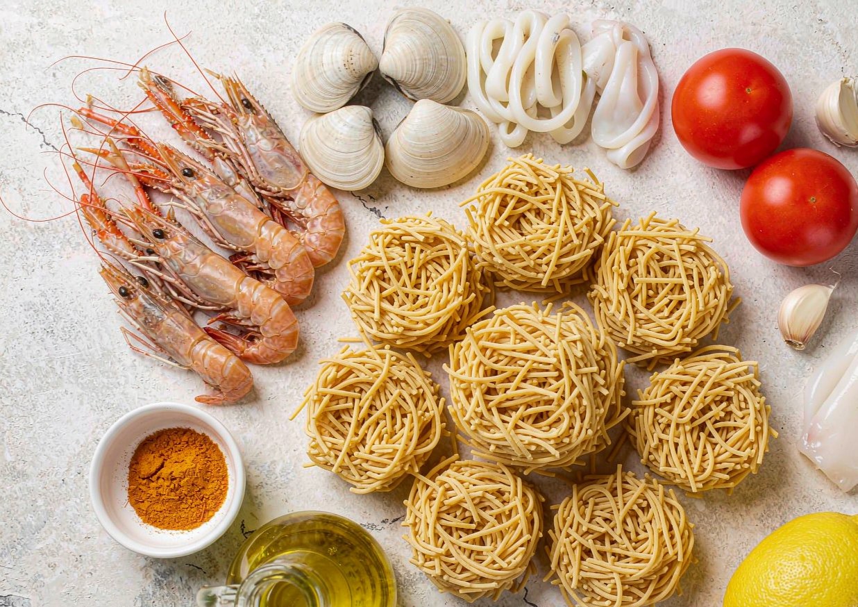 Overhead flat lay of fideua ingredients including fideus noodles, prawns, clams, squid, garlic, tomatoes and saffron
