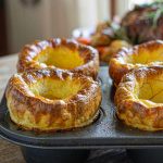 Four golden Yorkshire puddings risen tall above the rim of a metal tin with crisp uneven tops