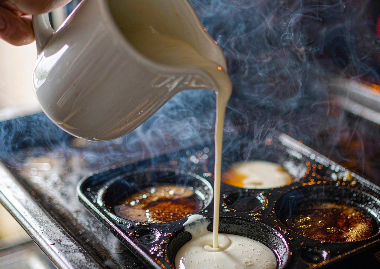 Batter being poured into hot beef dripping in a four-hole Yorkshire pudding tin in the oven